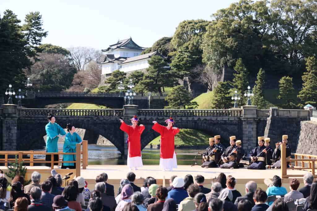 2 dancers in red, 2 dancers in blue on stage in front of bridge and Japanese palace