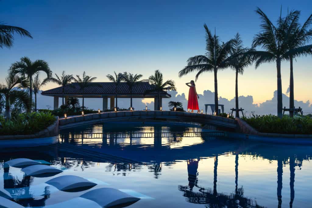 woman in red dress on edge of big pool in the evening among candlelight, with blue cloud backdrop.