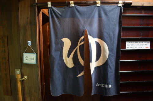 Noren bleu sur lequel est inscrit le caractère "yu", indiquant l'entrée de la salle de bains