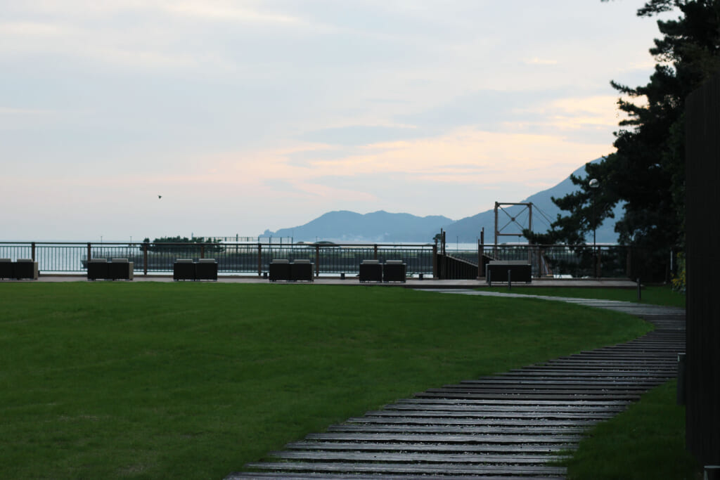 Au premier plan une pelouse est parcourue par un chemin de planches sur la gauche. Ce chemin mène à la terrace au bout de la pelouse qui se trouve face à la mer. Au loin on aperçoit des îles et des montagnes.