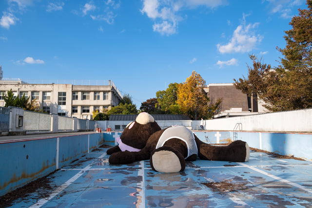Precious Okoyomon, Touching My Lil Tail Till the Sun Notices Me : un ours en peluche géant avec une petite culotte, couché dans une piscine d'école vide