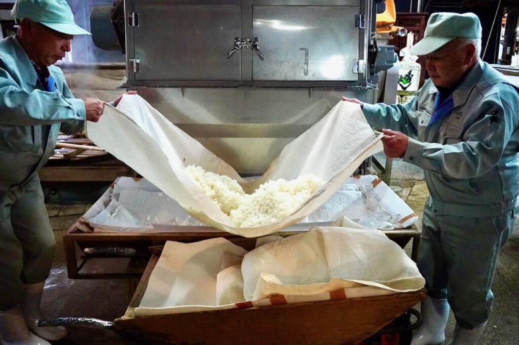 deux hommes mettent du riz cuit à la vapeur dans une brouette de la brasserie de saké Suzuki｜Hideyoshi